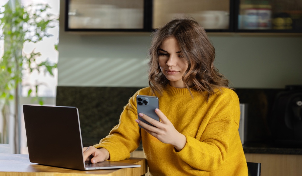 A girl in yellow sweater looking on a phone while working on a laptop