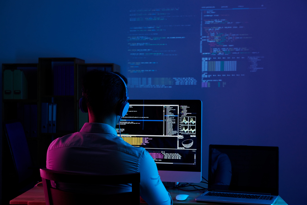 Software developer wearing headphones, working on multiple screens with code projected on the wall in a dimly lit office.